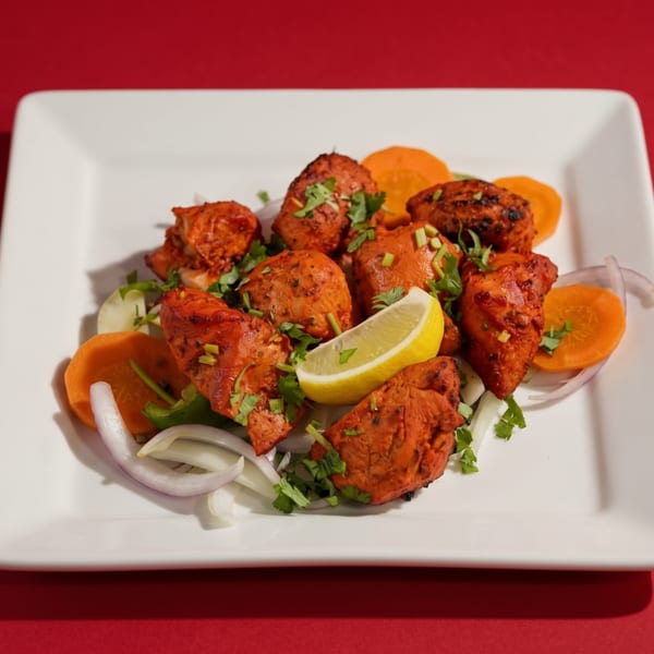 Tandoori chicken tikka with lemon, onions, carrots, and cilantro on a white plate.