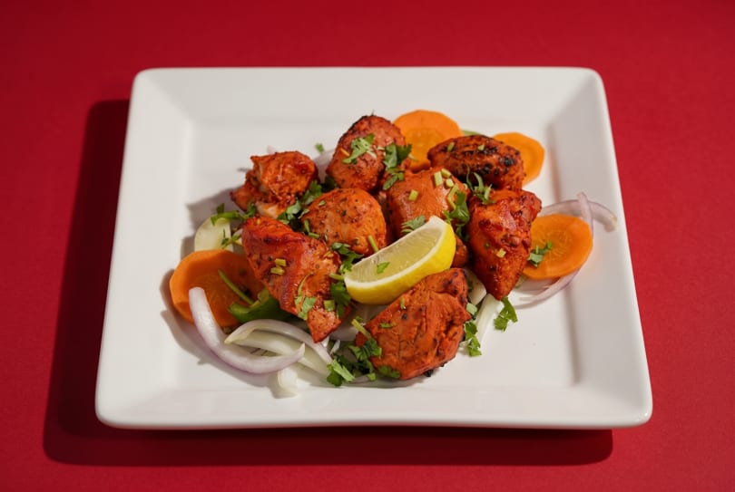 Tandoori chicken tikka with lemon, onions, carrots, and cilantro on a white plate.