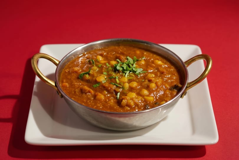 A metal bowl of chana masala, garnished with cilantro, sits on a white plate.