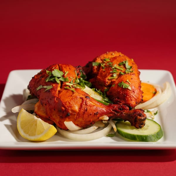 Two pieces of Tandoori chicken on a white plate with onions, cucumber, and lemon.