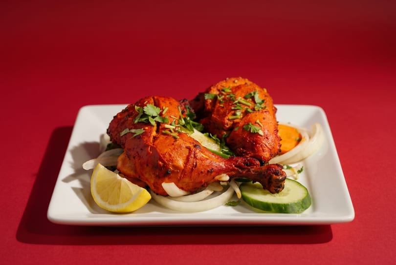 Two pieces of Tandoori chicken on a white plate with onions, cucumber, and lemon.