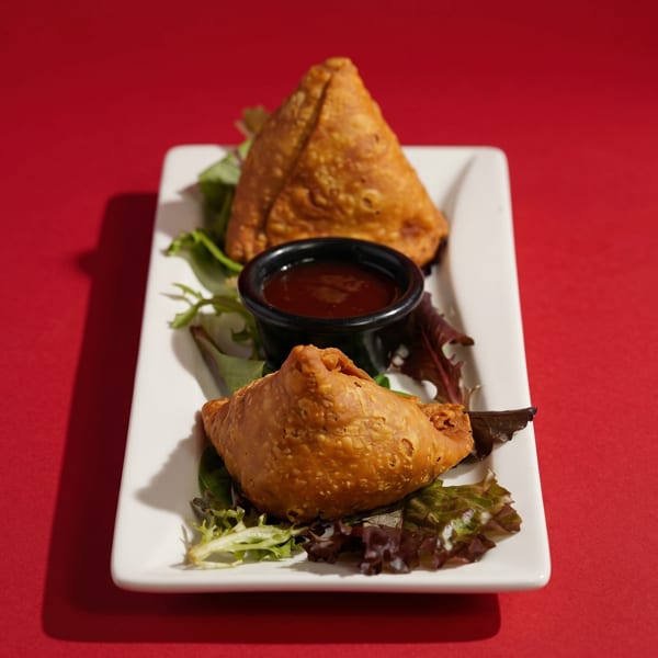 Two golden-brown samosas with a side of dipping sauce and lettuce on a white plate.