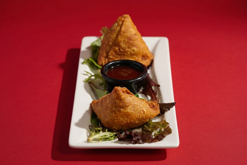 Two golden-brown samosas with a side of dipping sauce and lettuce on a white plate.