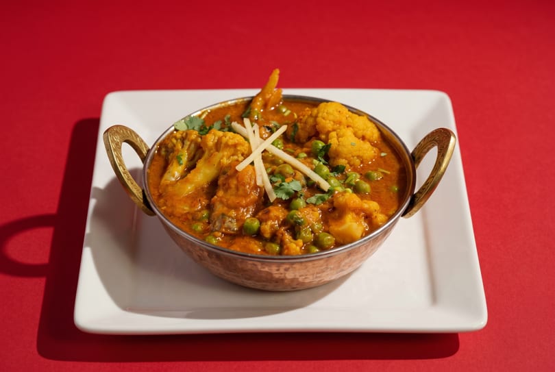 A copper bowl of Indian curry with cauliflower, peas, and ginger on a white plate.