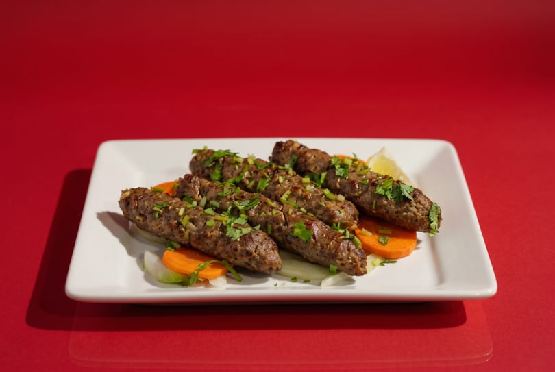 Five seekh kebabs garnished with cilantro and served with sliced carrots and onions.