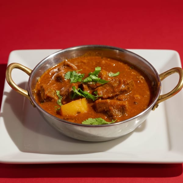 A metal karahi bowl filled with rich, orange-brown curry, garnished with fresh cilantro, on a white plate.