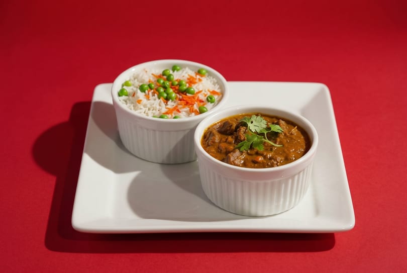 Two white ramekins on a rectangular plate, one with rice and peas, the other with beef curry.